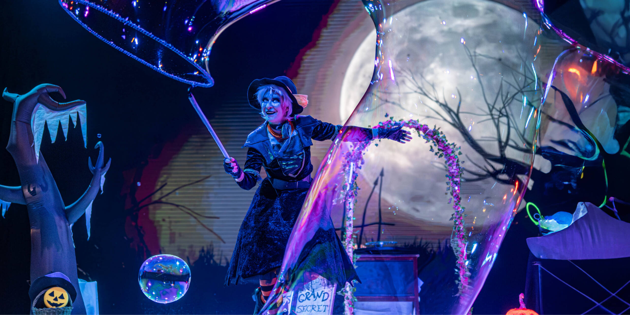Artista disfrazada de bruja creando una gran pompa de jabón en un espectáculo de Halloween con luna llena